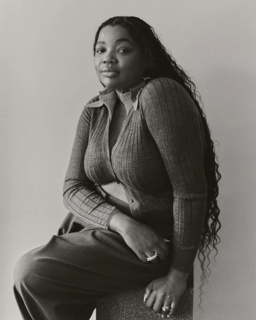Monochrome portrait of a Black woman with long dark wavy hair seated wearing a ribbed button-up cardigan and trousers.