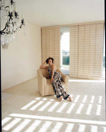 Woman in patterned jumpsuit seated in a beige armchair inside a sunlit room with a crystal chandelier and shuttered doors.