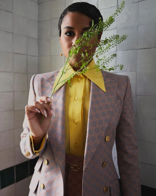 Stylish portrait of an adult woman wearing a Gucci blazer and yellow blouse, holding a leafy green plant in front of tiled wall.