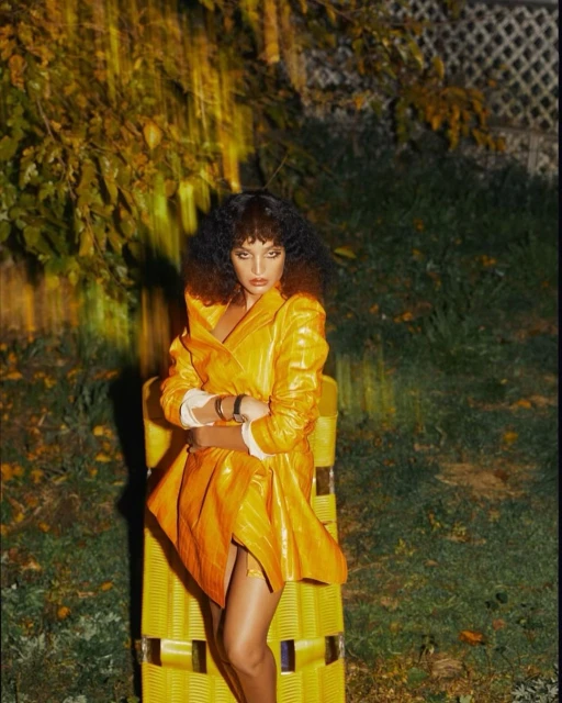 A woman with dark curly hair poses in a shiny yellow coat and short skirt on a yellow chair outdoors, surrounded by leafy greenery.