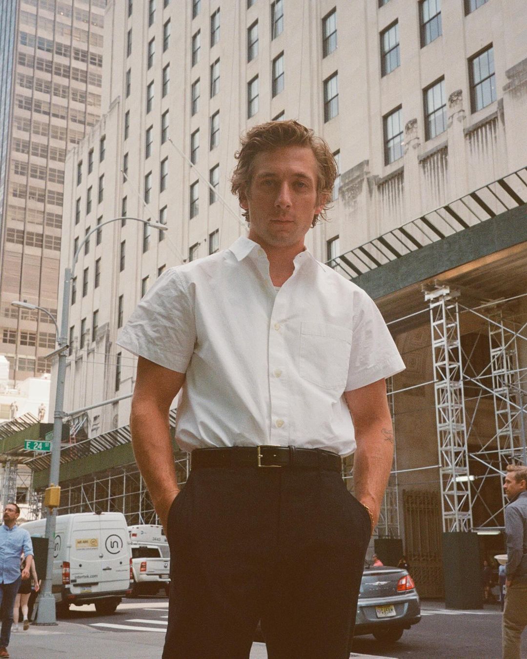 Adult male in white short-sleeve dress shirt, black pants with hands in pockets on a city street with buildings and pedestrians.