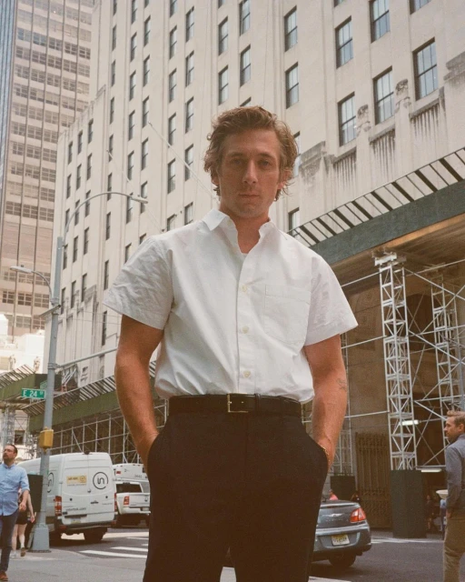 Adult male in white short-sleeve dress shirt, black pants with hands in pockets on a city street with buildings and pedestrians.