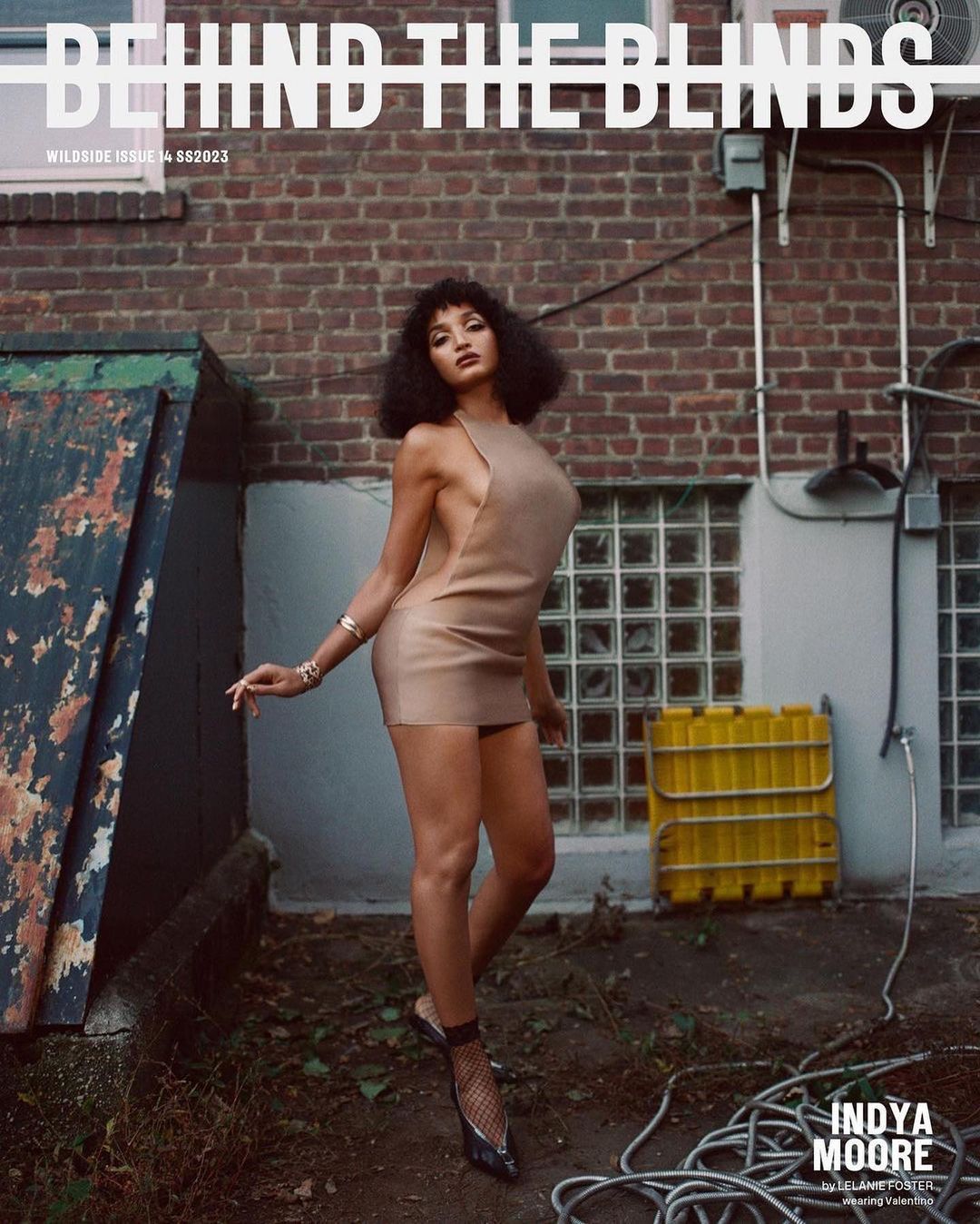 Indya Moore poses in front of a brick wall wearing a short dress and heels for a magazine cover titled 'Behind the Blinds'.