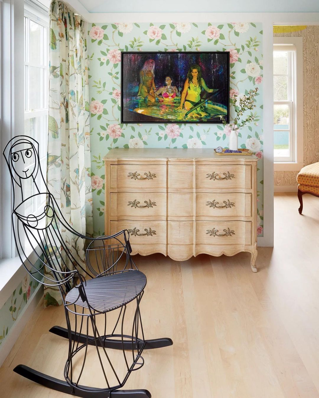 Living room interior with a black wire rocking chair, a light wood dresser, and a painting of women above the dresser.