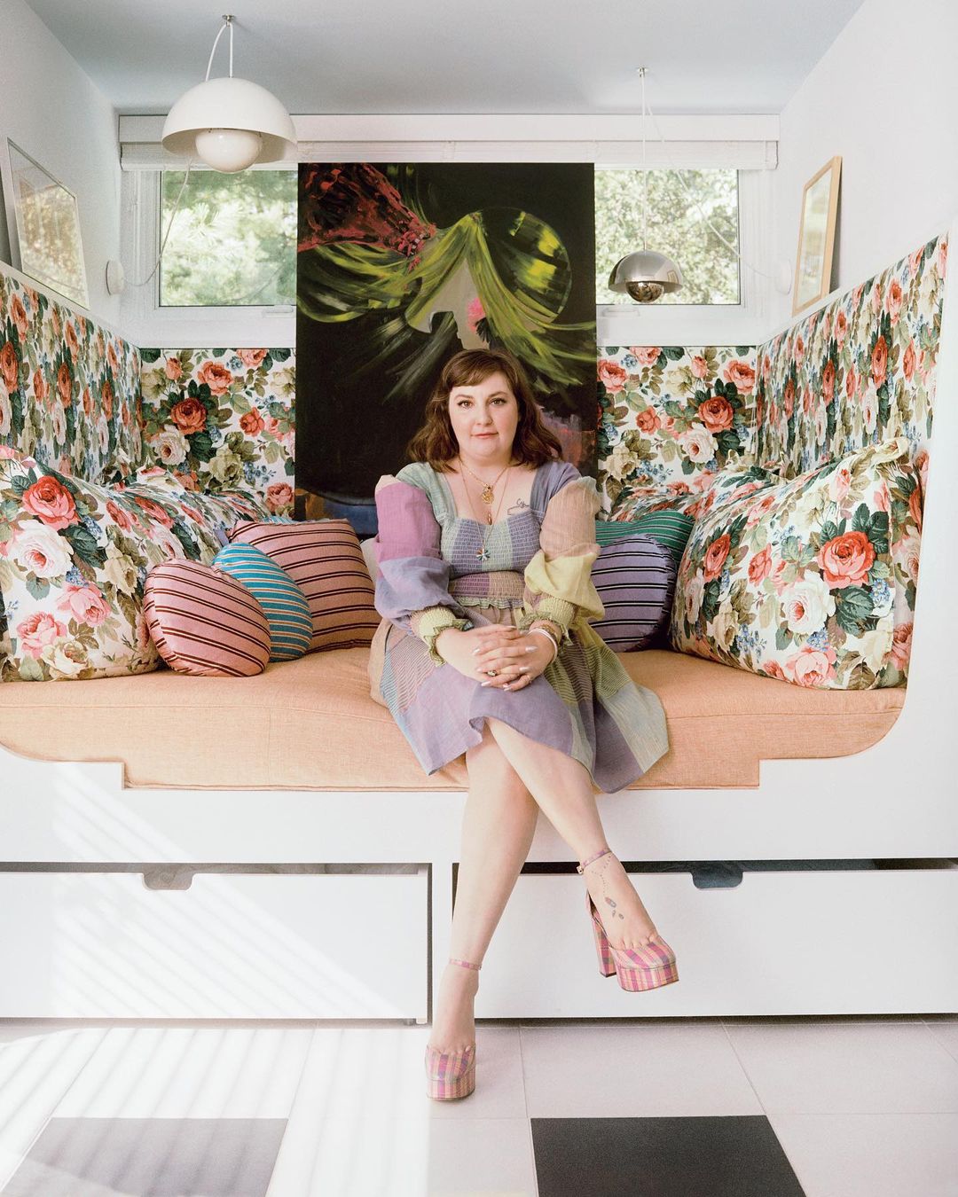 Adult female seated on a floral sofa in a brightly lit interior, wearing a multicolored patchwork dress and pink platform heels.
