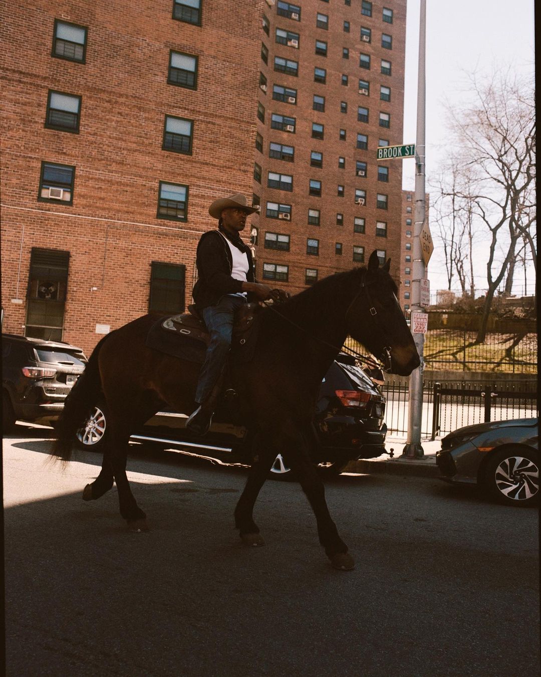 A person wearing a cowboy hat riding a horse on a city street with brick buildings and parked cars on either side.