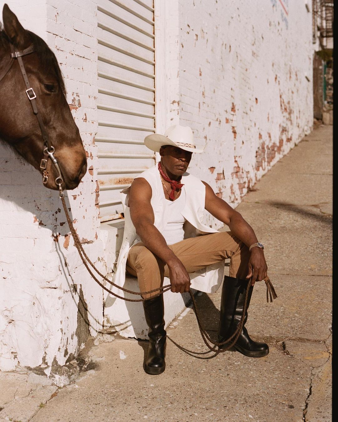 African-American man in cowboy hat and vest seated next to a horse against a white brick wall in an urban setting, holding a whip.