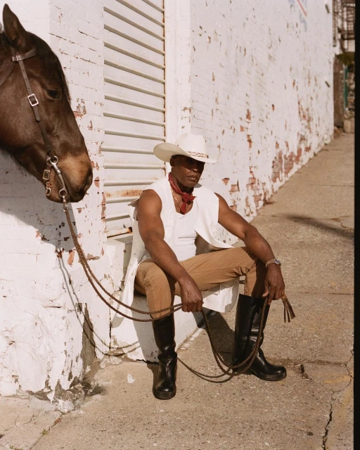 African-American man in cowboy hat and vest seated next to a horse against a white brick wall in an urban setting, holding a whip.