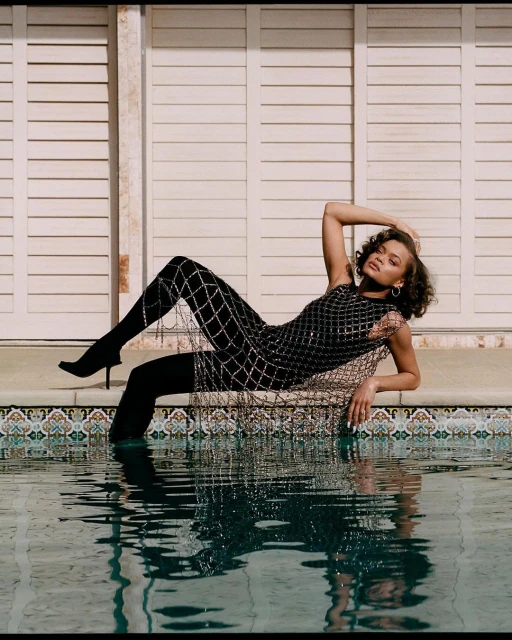 Woman in a black sequin dress and leggings poses in a pool with hands behind head. Pool is surrounded by a tiled patio and white siding.
