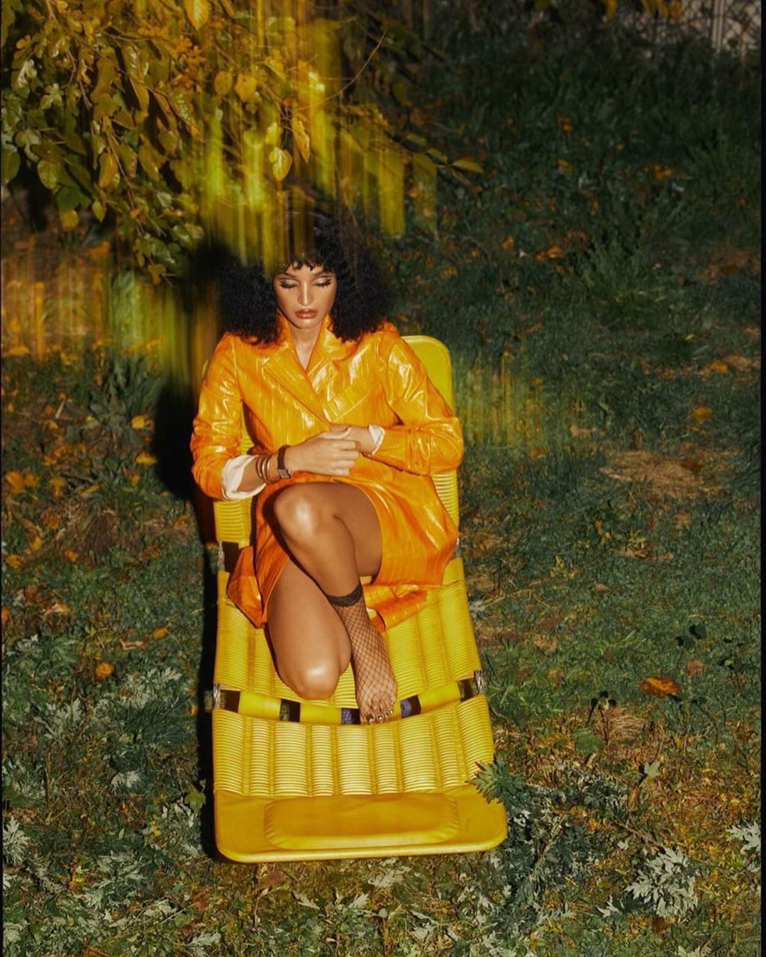 Woman in a yellow vinyl outfit sits in a matching yellow deck chair surrounded by green foliage, creating a summery outdoor scene.