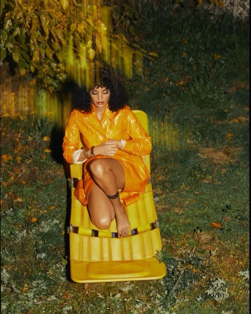 Woman in a yellow vinyl outfit sits in a matching yellow deck chair surrounded by green foliage, creating a summery outdoor scene.