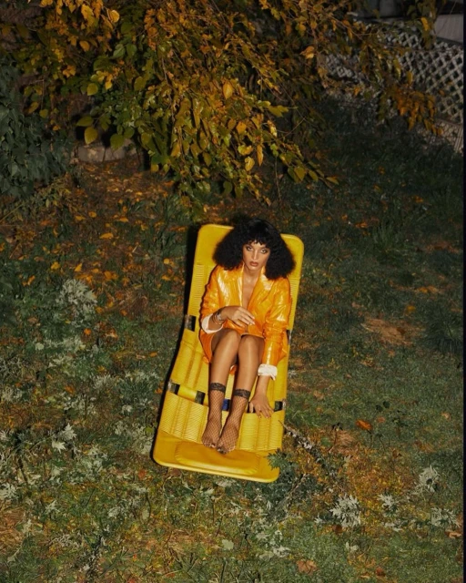 A woman with curly hair lounges on a yellow raft on grassy ground under a leafy tree, wearing a shiny orange jacket and fishnet stockings.