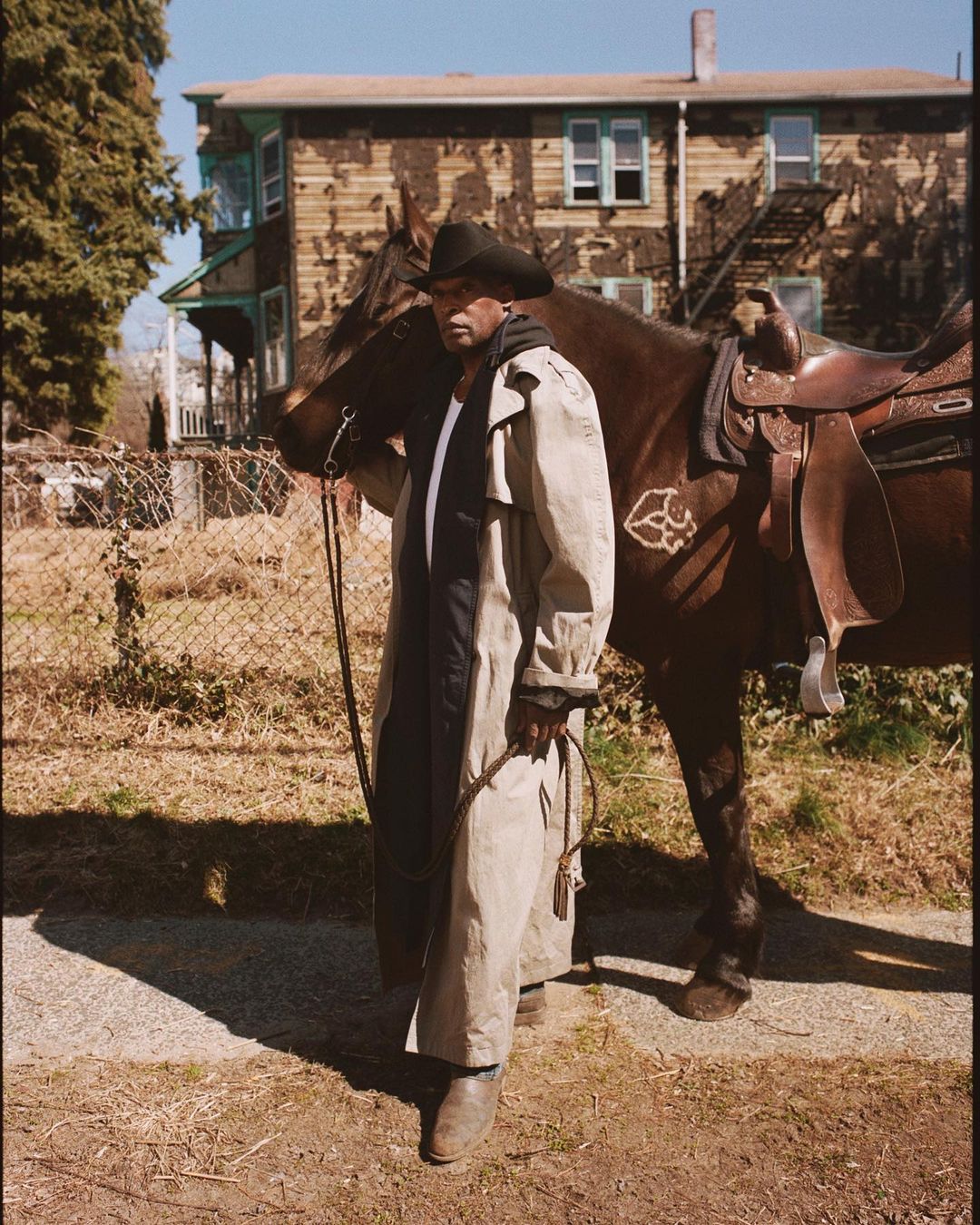 An African-American man in cowboy hat and tan coat stands next to a saddled horse on a sunny day in front of a weathered building.