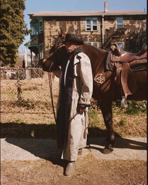 An African-American man in cowboy hat and tan coat stands next to a saddled horse on a sunny day in front of a weathered building.