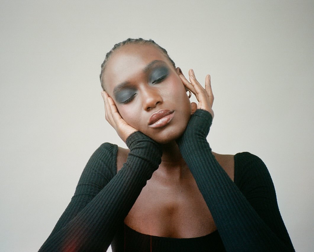Close-up of an African-American woman with eyes closed, wearing dark eye shadow and a long-sleeved black shirt, set against a neutral backdrop.