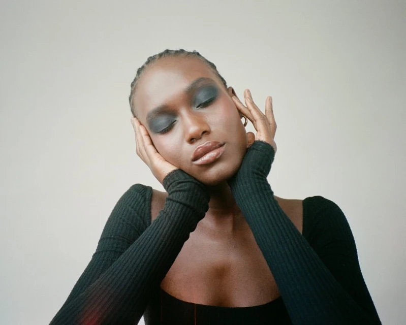 Close-up of an African-American woman with eyes closed, wearing dark eye shadow and a long-sleeved black shirt, set against a neutral backdrop.