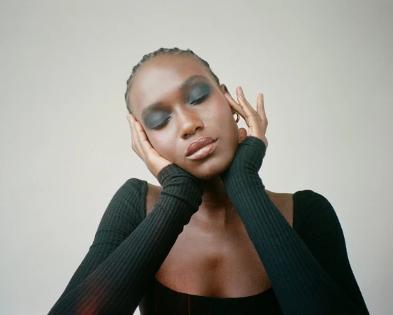 Close-up of an African-American woman with eyes closed, wearing dark eye shadow and a long-sleeved black shirt, set against a neutral backdrop.