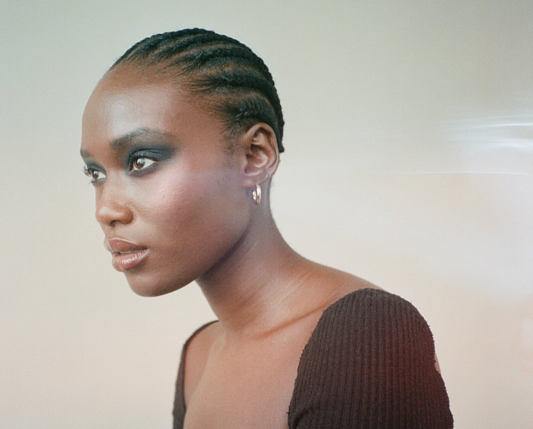 Close-up studio shot of a Black female model with cornrow braids, wearing smoky eyeshadow and a dark brown top against an off-white background.