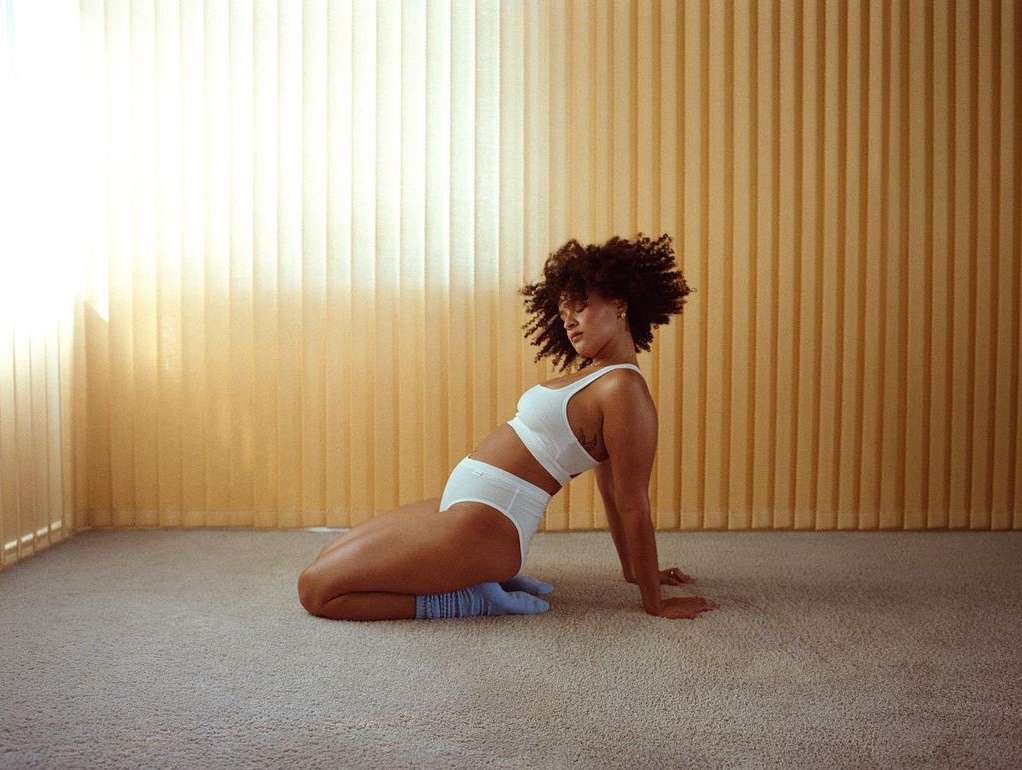 Woman with curly hair in white sports bra and shorts kneels indoors on carpet, looking off camera near tan blinds and light.