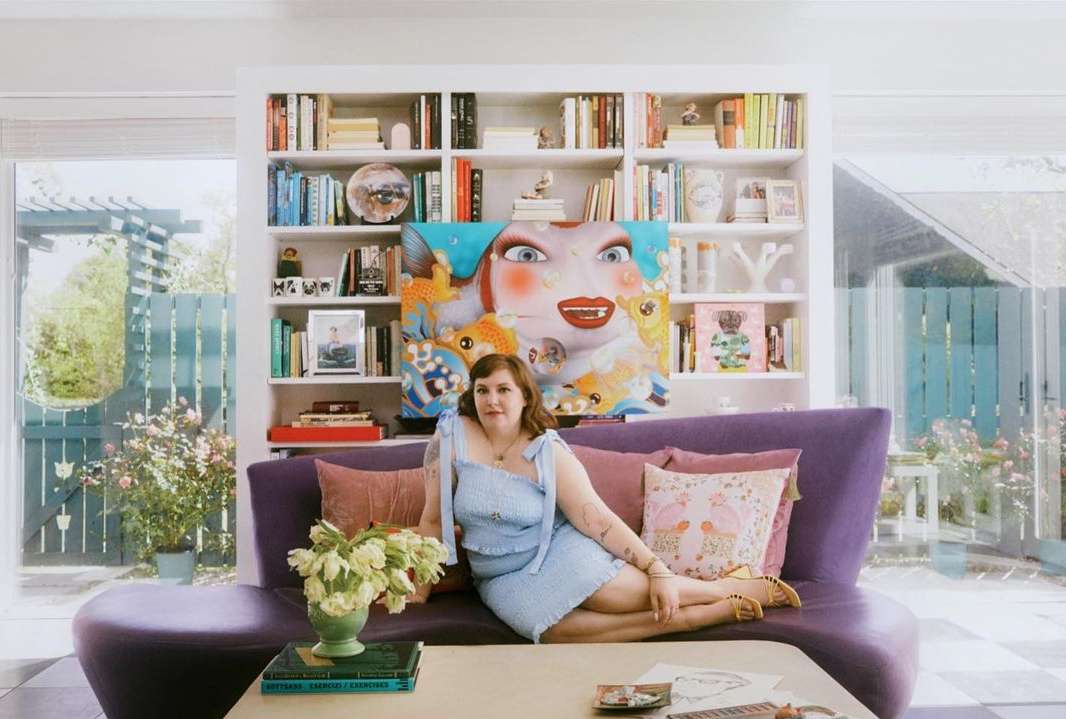 Woman in blue dress reclines on purple sofa in a sunlit indoor setting with a bookshelf and art. Flowers and books on table.