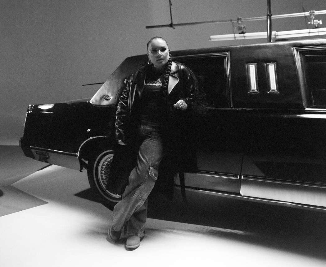 Black and white portrait of a woman leaning against a limo. She wears a long leather coat, t-shirt, and jeans, with a braid over her shoulder.