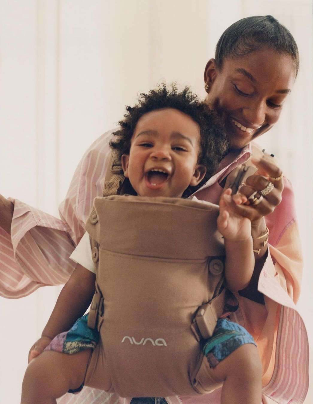 Happy baby smiles while being held in a tan-colored Nuna baby carrier by an adult female with short hair in a bright indoor space.
