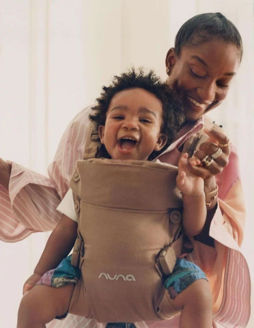 Happy baby smiles while being held in a tan-colored Nuna baby carrier by an adult female with short hair in a bright indoor space.