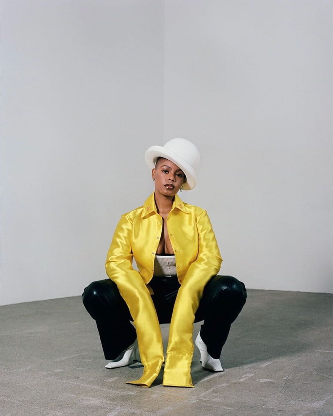 Woman in yellow jacket, black pants and white hat squats in a photo studio, gazing at the camera with a neutral expression.