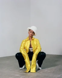 Black woman squatting, wearing a yellow satin button-down shirt with long sleeves, black pants, white derby hat, and white heels.