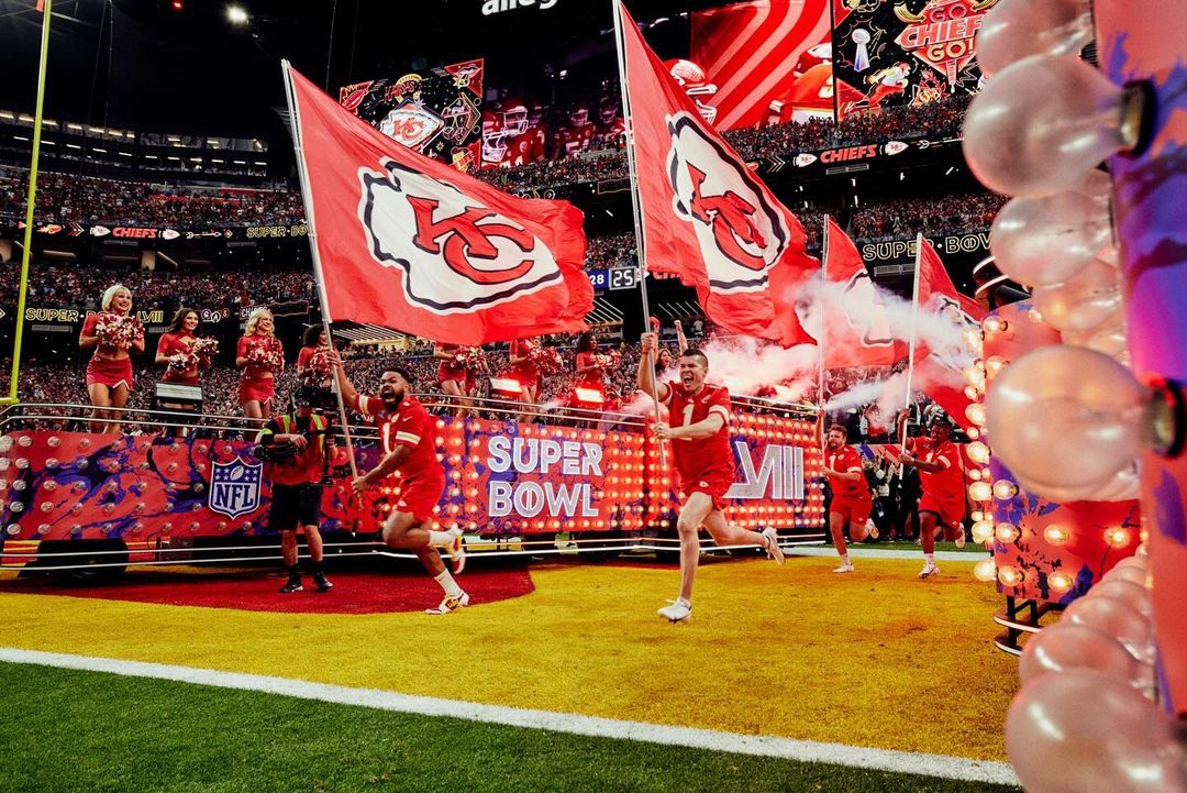 Kansas City Chiefs players run onto a football field holding team flags during Super Bowl LVII, surrounded by cheerleaders and fans.