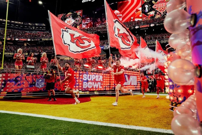 Kansas City Chiefs players run onto a football field holding team flags during Super Bowl LVII, surrounded by cheerleaders and fans.