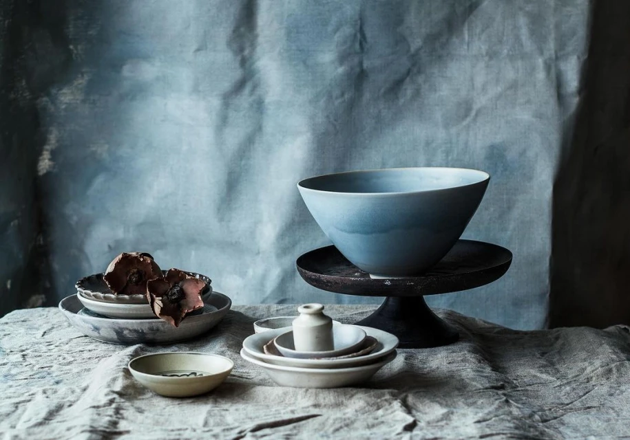 Still life composition featuring a large blue bowl, plates, pottery, and dried flowers on a textured grey cloth against a mottled backdrop.