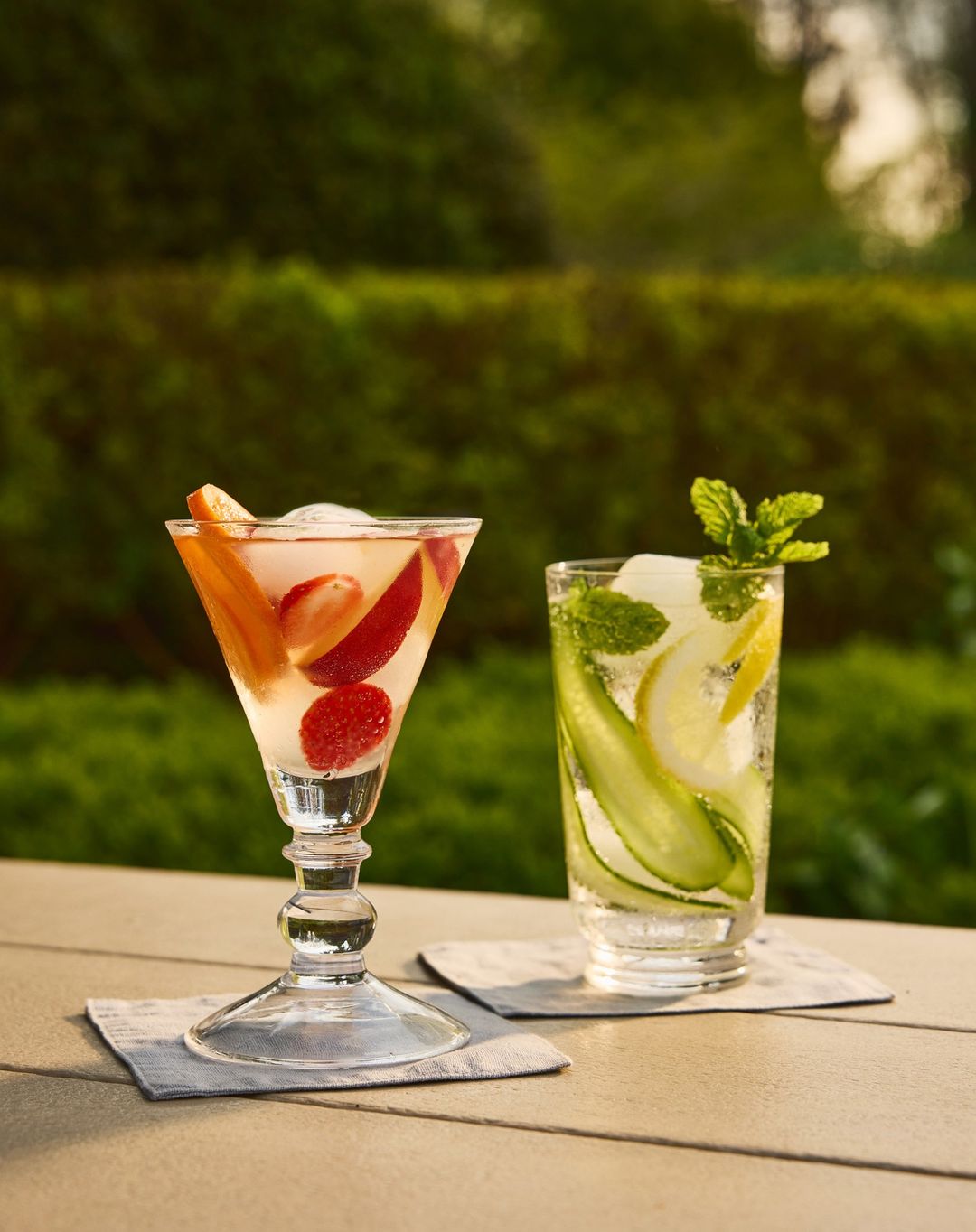 Two refreshing drinks on a table, one in a stemmed glass with fruit, the other in a clear glass with cucumber and mint, outdoors.