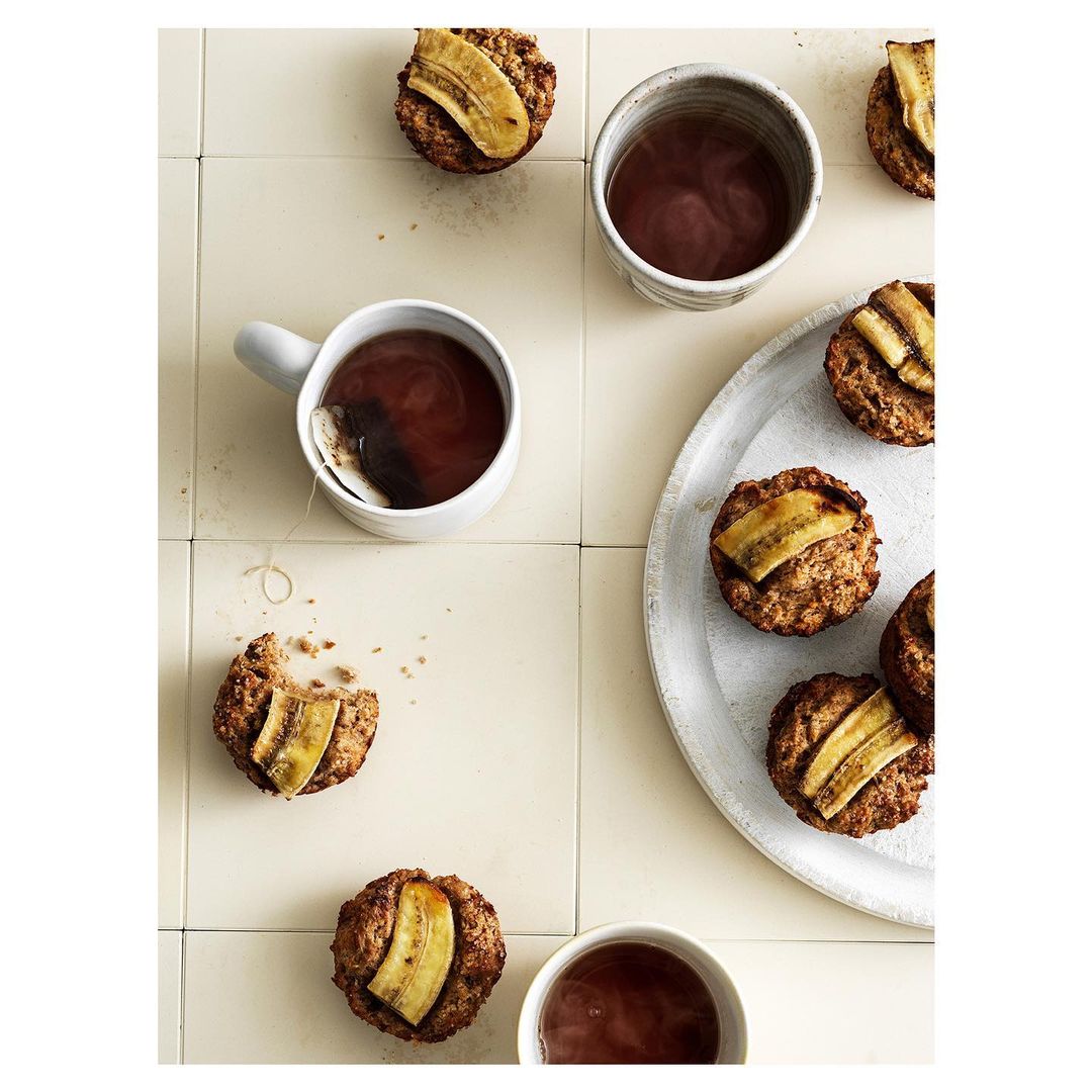 Overhead shot of banana muffins on a white plate, tea in white mugs, and scattered muffins, set against a light tile background.