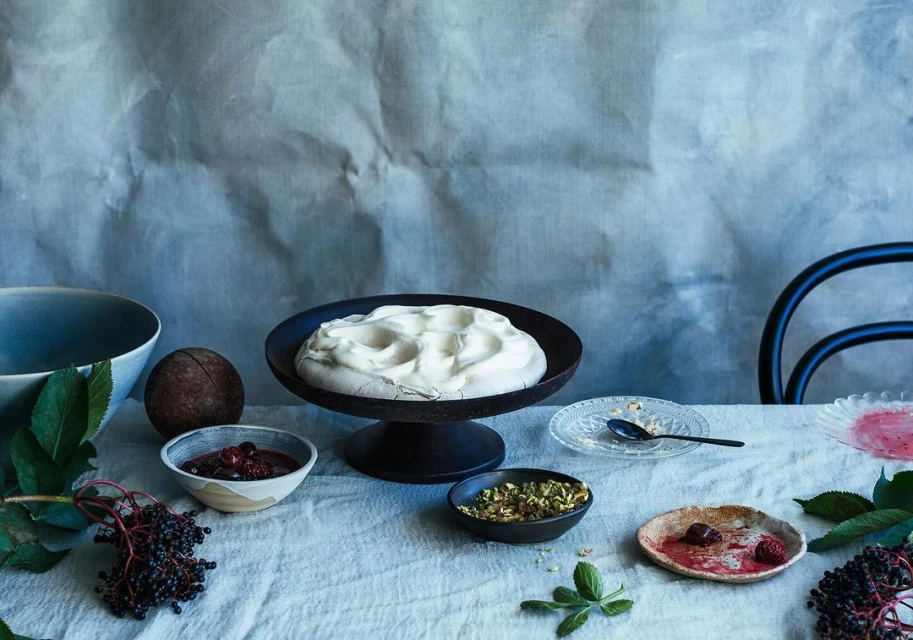 Overhead shot of a dessert stand with a meringues, bowls of fruit and nuts, and stems of elderberries on a white linen tablecloth.