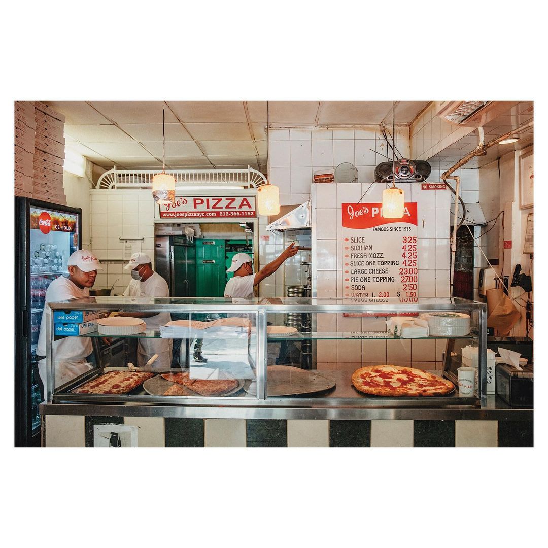 Inside a bustling pizzeria, three adult male pizza makers prepare dishes behind a glass display case filled with pizzas and desserts.