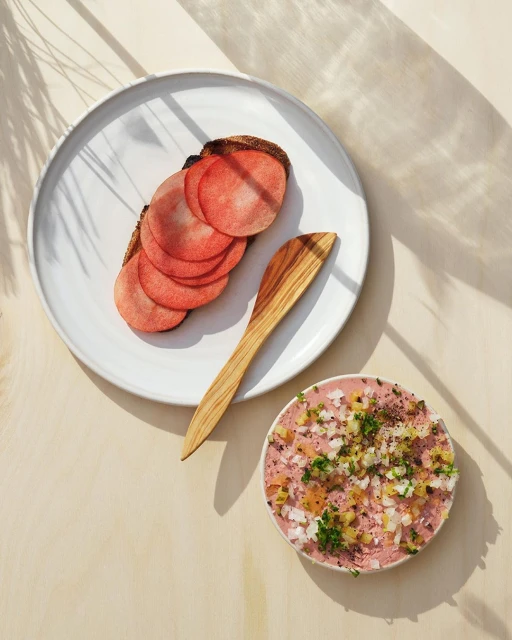 Overhead shot of a white plate with sausage slices on toast, next to a small bowl of sausage spread, with light and shadows creating a minimalist food still life.
