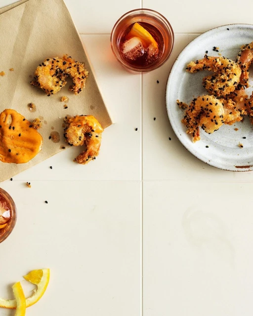 Overhead shot of crispy shrimp with black sesame seeds, sauce, drink, and orange slices on a white tiled surface for food photography.