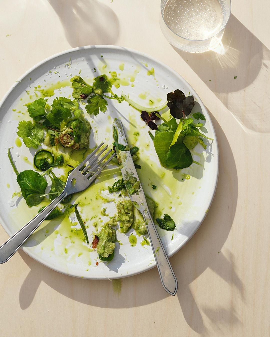 Overhead view of leftovers on a white plate with a fork, knife, and glass of water, set on a neutral-toned wooden table, food photography.