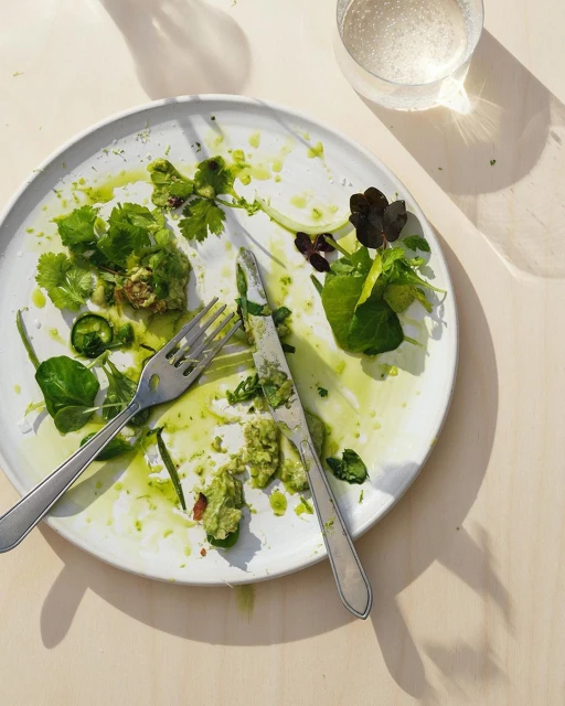 Overhead view of leftovers on a white plate with a fork, knife, and glass of water, set on a neutral-toned wooden table, food photography.