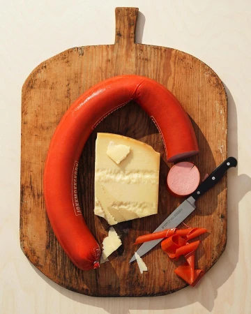 Overhead shot of sausage, cheese, and a knife on a wooden cutting board. Food photography for food and beverage industry concept.