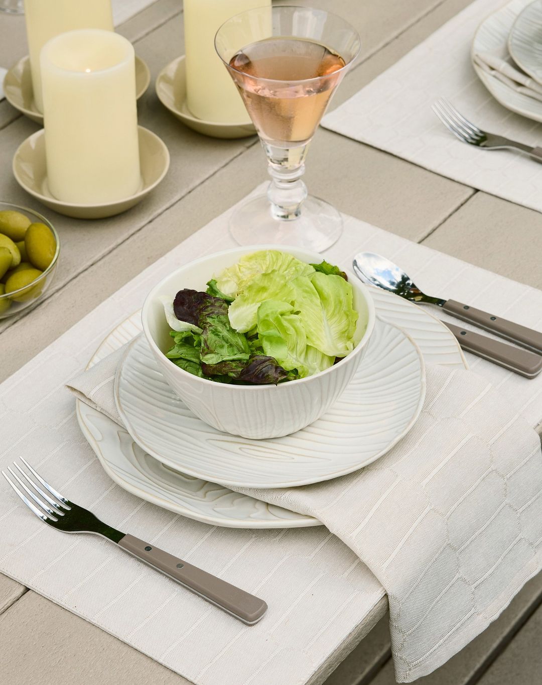 Overhead view of a place setting on a wooden table, featuring a green salad, elegant plates, silverware, candles, and a pink drink.