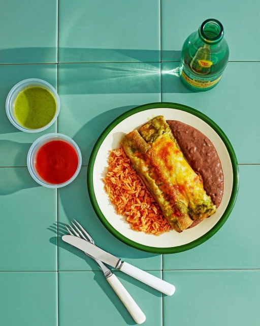 Overhead shot of enchiladas with rice and beans on a white plate, along with salsa and a bottle on a turquoise tile surface.