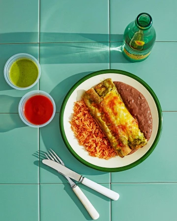 Overhead shot of enchiladas with rice and beans on a white plate, along with salsa and a bottle on a turquoise tile surface.
