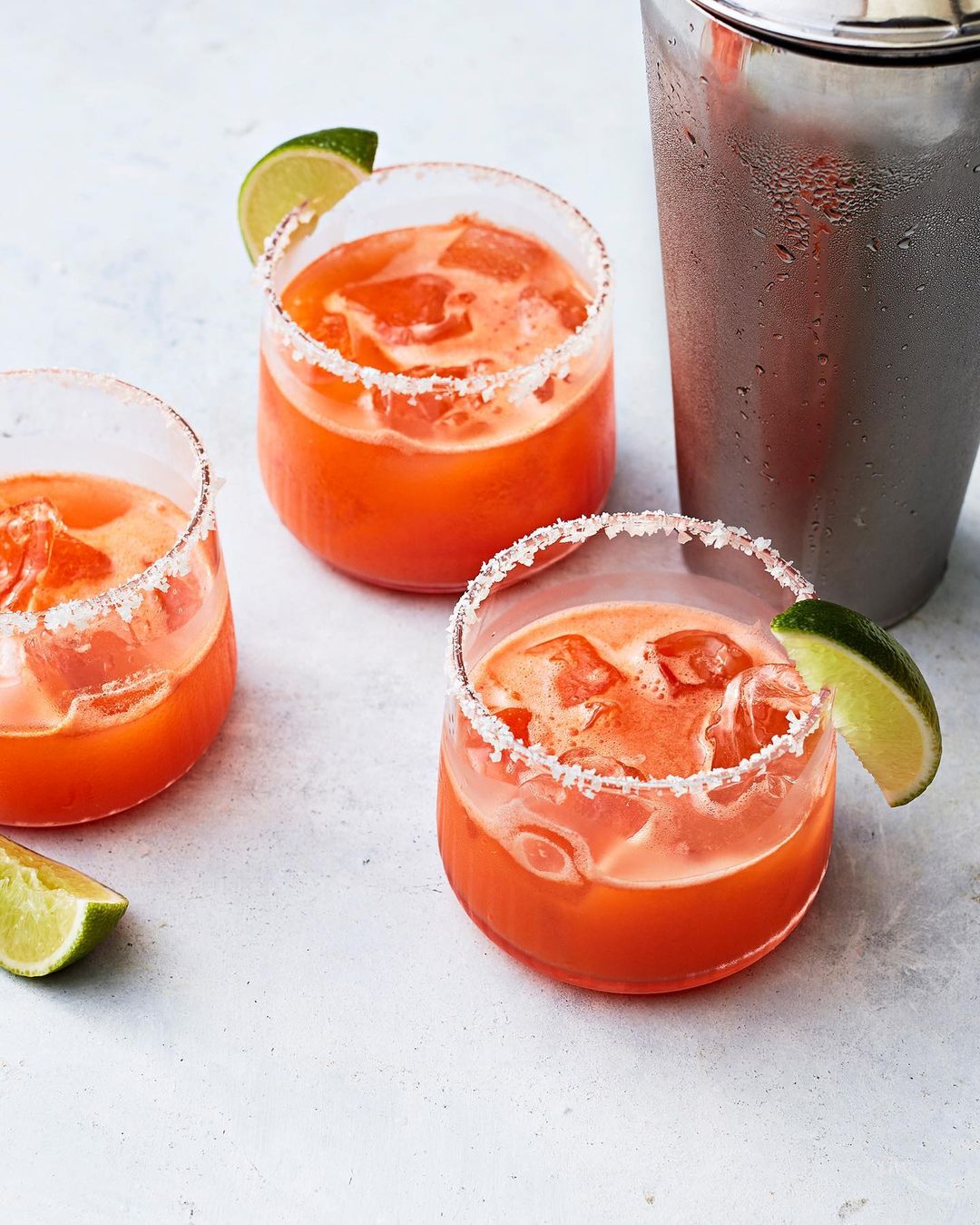 Three salt-rimmed glasses filled with orange cocktails, garnished with lime wedges. A metal cocktail shaker stands in the background.
