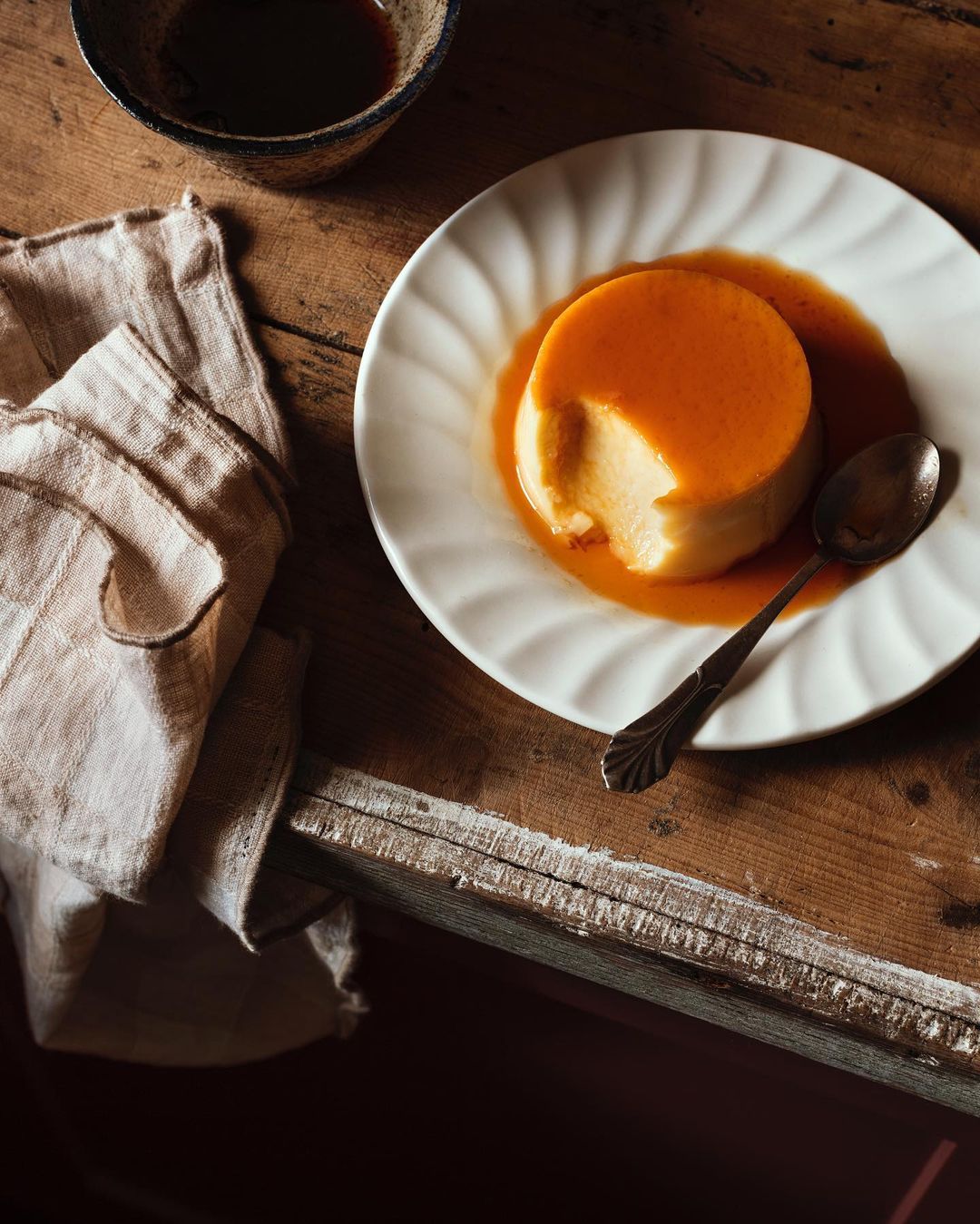 A caramel flan dessert on a white plate with a spoon, next to a linen cloth and small bowl on a rustic wooden table in warm light.