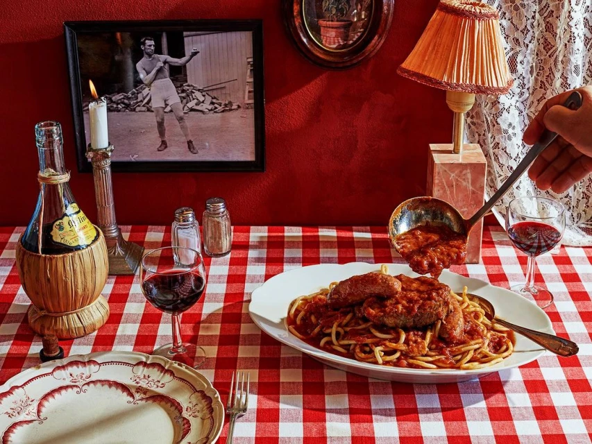 Spaghetti with tomato sauce and meat on a red and white checkered tablecloth, with wine and a photo of a boxer on the wall.