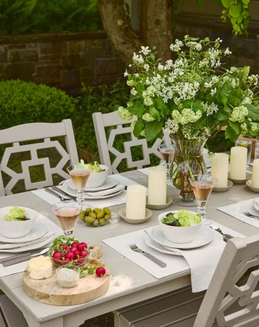 Outdoor dining table set with plates, salad bowls, glasses, candles, and white floral arrangement. Garden and trees in the background.