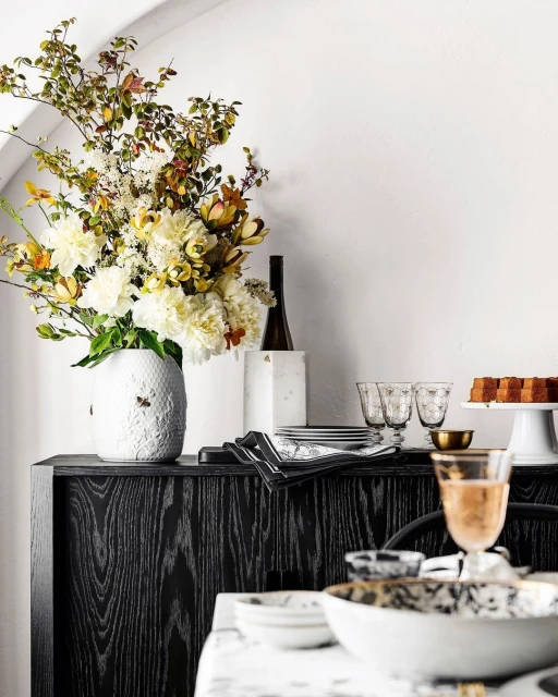 Stylized table setting featuring a floral arrangement, glassware, plates, and linens on a black sideboard against a white wall backdrop.