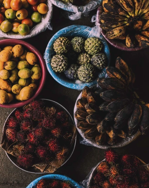 Overhead shot showcases a colorful assortment of tropical fruits, including bananas, lychees, mangoes, sugar-apples, and tomatoes arranged in circular baskets.